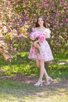 Adorable girl, stands with a large bouquet of peonies, near pink blooming garden, on a sunny day. Copy space. Vertical