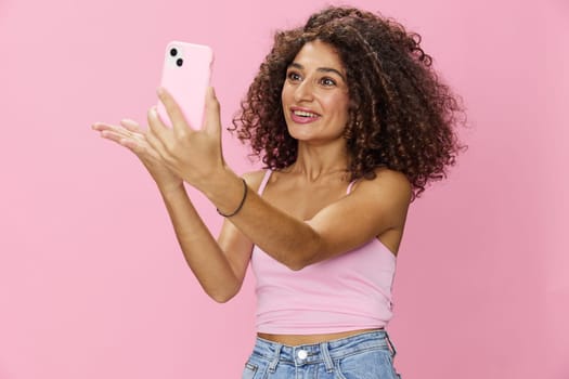 Woman blogger holding phone in hand video call, with curly hair in pink top and jeans poses on pink background, copy space, technology and social media, online. High quality photo