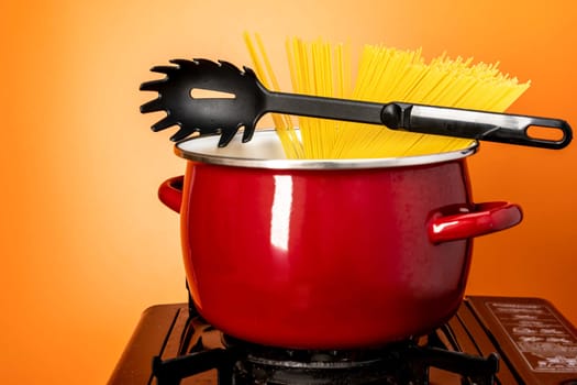we cook ourselves. spaghetti in a saucepan. a beautiful red saucepan with spaghetti and skimmer. cooking spaghetti in a saucepan on a gas stove. homemade Italian-style dinner. kitchen saucepan on a orange background