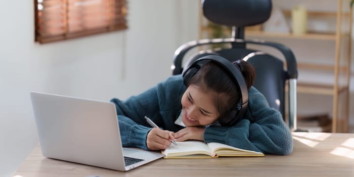 Online education, e-learning. Asian woman in stylish casual clothes, studying using a laptop, listening to online lecture, taking notes, online study at home.