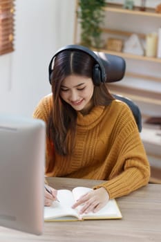 Online education, e-learning. Asian woman in stylish casual clothes, studying using a computer, listening to online lecture, taking notes, online study at home.