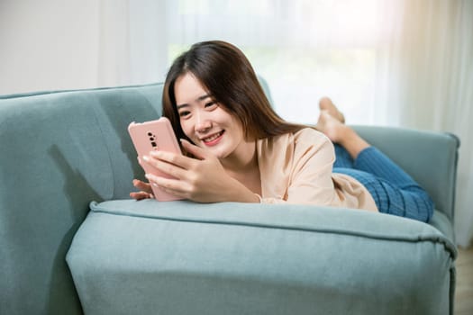 Smiling woman relaxed social online communication on smartphone, Happy Asian female using smart mobile phone lying on sofa in living room room at house, technology gadget online application