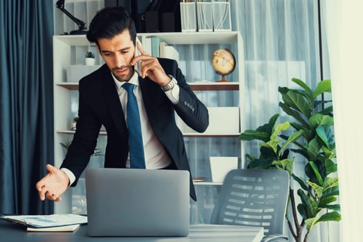 Diligent businessman busy talking on the phone call with clients while working with laptop in his office as concept of modern hardworking office worker lifestyle with mobile phone. Fervent