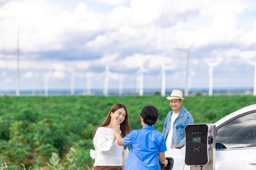 Concept of progressive happy family enjoying their time at wind farm with electric vehicle. Electric vehicle driven by clean renewable energy from wind turbine generator for charging station.