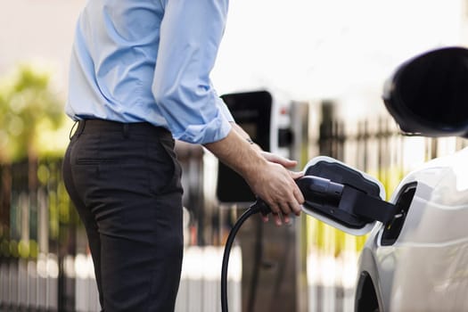 Closeup progressive man holding EV charger plug from public charging station for electric vehicle with background of residential building as concept eco-friendly sustainability energy car concept.