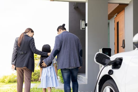 Progressive young parents and daughter with electric vehicle and home charging station. Green and clean energy from electric vehicles for healthy environment. Eco power from renewable source at home.