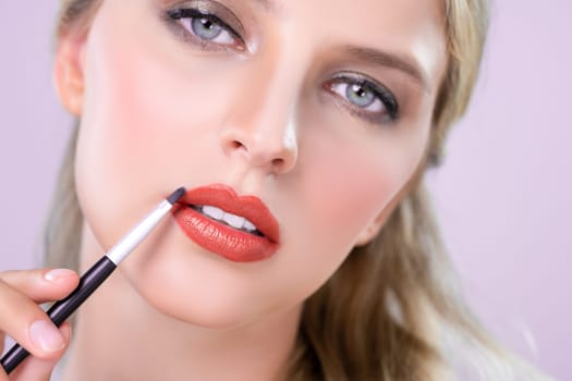 Closeup beautiful young woman with flawless healthy skin and natural makeup putting alluring fashion glossy red lipstick on her lip with lip brush in pink isolated background.