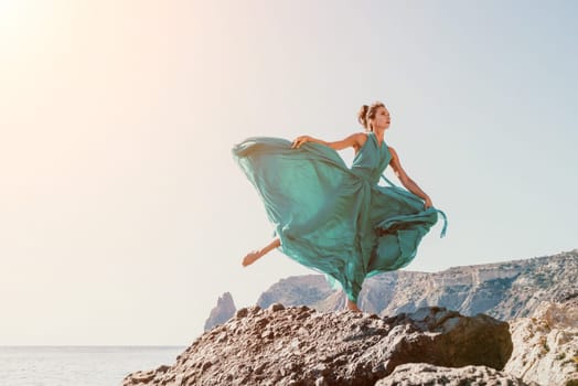 Side view a Young beautiful sensual woman in a red long dress posing on a rock high above the sea during sunrise. Girl on the nature on blue sky background. Fashion photo.