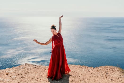 Side view a Young beautiful sensual woman in a red long dress posing on a rock high above the sea during sunrise. Girl on the nature on blue sky background. Fashion photo.