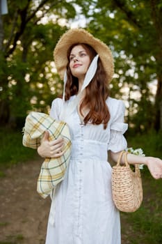 a happy woman in a light summer dress stands in nature with a wicker hat, a plaid and a basket with daisies. High quality photo