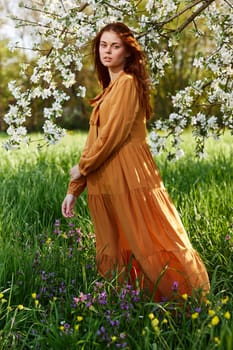 a bright vertical photo of an attractive woman in a long orange dress standing next to a flowering tree in sunny, warm weather, looking pleasantly into the camera. High quality photo