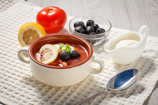 Soup saltwort with meat, smoked sausages, potatoes, tomatoes, marinated pickled cucumber, lemon, black olives and sour cream, ceramic soup bowl with ingredients on white kitchen towel.