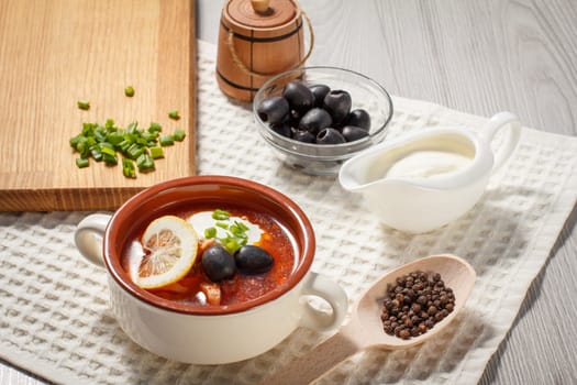 Soup saltwort with sausages, potatoes, tomatoes, marinated pickled cucumber, lemon, black olives and sour cream, ceramic soup bowl with ingredients, wooden spoon with peppercorns, cutting board.