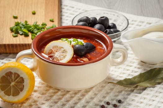 Soup saltwort with meat, smoked sausages, potatoes, tomatoes, marinated pickled cucumber, lemon, black olives and sour cream in ceramic soup bowl with ingredients.