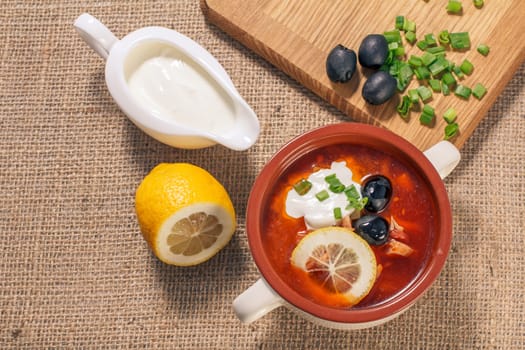 Soup saltwort with meat, smoked sausages, potatoes, tomatoes, marinated pickled cucumber, lemon, black olives and sour cream in ceramic soup bowl with ingredients. Top view