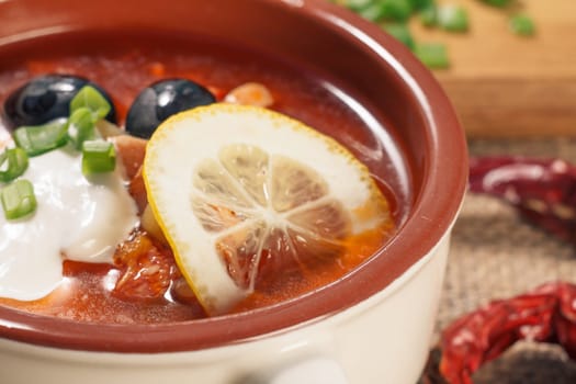 Close-up soup saltwort with meat, smoked sausages, potatoes, tomatoes, marinated pickled cucumber, lemon, black olives and sour cream in ceramic soup bowl, dried red peppers. Selective focus.