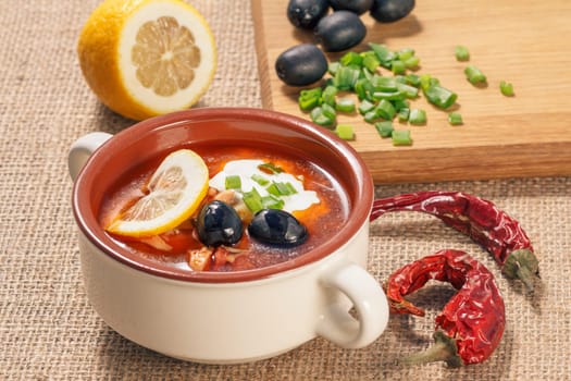 Soup saltwort with meat, smoked sausages, potatoes, tomatoes, marinated pickled cucumber, lemon, black olives and sour cream in ceramic soup bowl, dried red peppers on sackcloth.