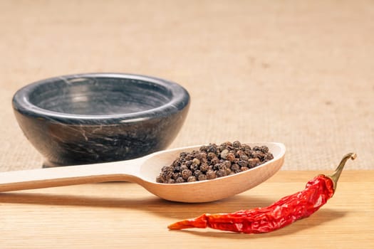 Wooden spoon with black peppercorns, dried red pepper on cutting board and stone mortar for grinding pepper in the background.