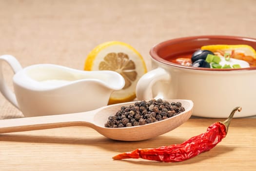 Wooden spoon with black peppercorn, dried red pepper on cutting board and ceramic soup bowl with saltwort, sauceboat with sour cream and cut lemon in the background.
