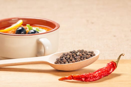 Wooden spoon with black peppercorn, dried red pepper on cutting board and ceramic soup bowl with saltwort in the background.