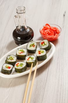 Sushi rolls with rice, pieces of avocado, cucumber, red bell pepper and lettuce leaves on ceramic plate, chopsticks, glass bottle with soy sauce and pickled ginger in a bowl. Vegetarian food.