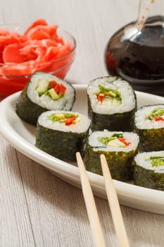 Sushi rolls with rice, pieces of avocado, cucumber, red bell pepper and lettuce leaves on ceramic plate, chopsticks, glass bottle with soy sauce and pickled ginger in a bowl. Vegetarian food.