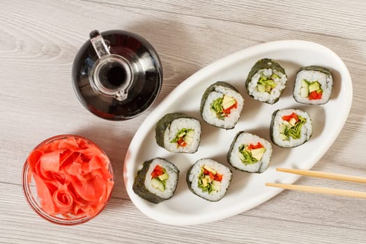 Sushi rolls with rice, pieces of avocado, cucumber, red bell pepper and lettuce leaves on ceramic plate, chopsticks, glass bottle with soy sauce and pickled ginger in a bowl. Top view