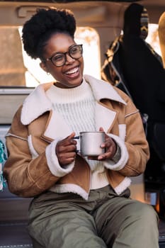 young black woman laughing happy at sunset sitting in a camper van with a cup of coffee in her hand, concept of van life and weekend getaway