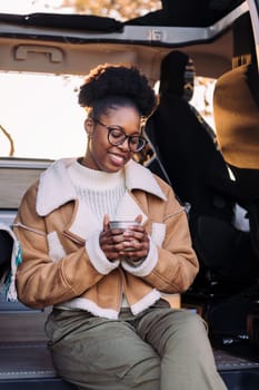 young african woman smiling happy at sunset relaxing in a camper van with a cup of a hot drink her hand, concept of van life and weekend getaway
