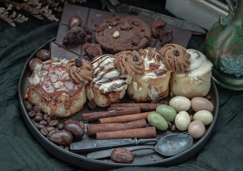 Assorted Cinnamon Rolls, Dark chocolate bars, cookies, pieces and balls served with black iron cutlery in black ceramic plate on dark background. Selective Focus.
