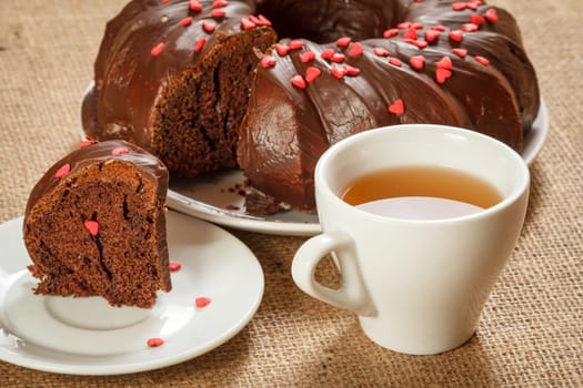 Slice of delicious homemade chocolate cake decorated with small hearts made of caramel on saucer and a cup of tea on table with sackcloth.