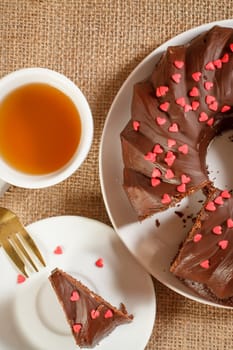 Homemade chocolate cake decorated with small hearts made of caramel on plate, cup of tea, slice of cake on saucer and fork on the table with sackcloth. Top view.