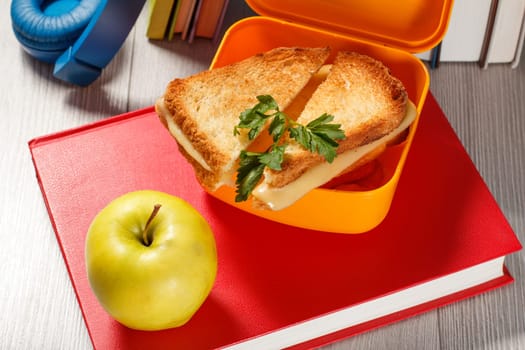 Yellow lunch box with toasted slices of bread, cheese and green parsley, green apple, book and headphones on the background. School breakfast.