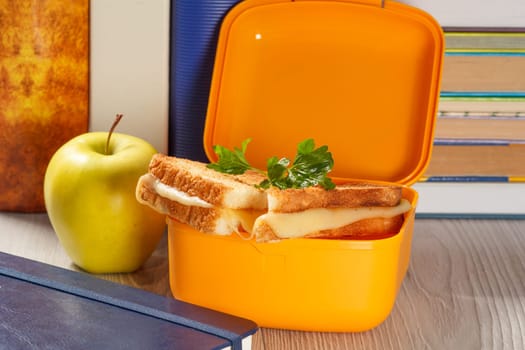 Yellow lunch box with toasted slices of bread, cheese and green parsley, green apple and hardback books on the background. School breakfast.