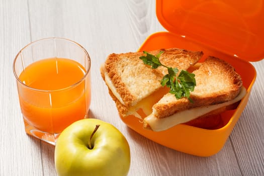 Yellow lunch box with toasted slices of bread, cheese and green parsley, green apple and glass of orange juice on wooden desk. School breakfast.