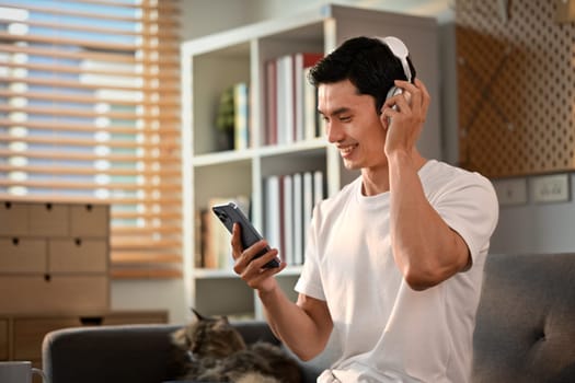 Happy Asian man enjoy favorite songs in headphones and using smartphone on couch. Technology and lifestyle concept.
