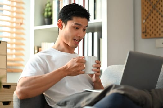 Handsome Asian man making video call on laptop sitting on couch at home. Technology, communicating and lifestyle.