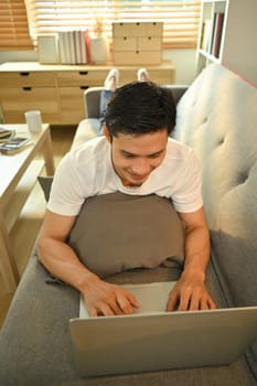 Relaxed asian man lying on couch and using laptop, surfing internet, working online, communication in social media.