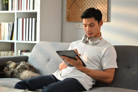 Image of asian man in casual clothes sitting on couch with cat and working on digital tablet. People, technology and lifestyle.