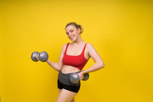 Beautiful model in sportswear with dumbbells looks at camera on yellow, red and dark background