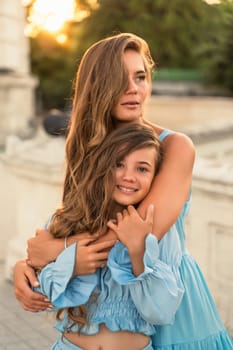 Mother aughter sunset. in blue dresses with flowing long hair against the backdrop of sunset. The woman hugs and presses the girl to her