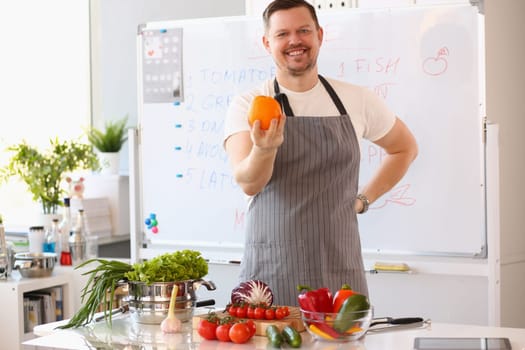 Male cook teacher in apron holds an orange and fresh vegetables and fruits on table. Culinary training and dietary nutrition