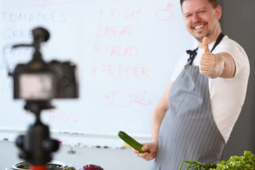 Laughing vlogger holding cucumber penis and filming video on camera. Joking chef holding thumbs up and cucumber