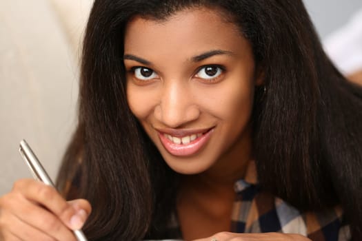 Young black woman writes in a magazine while lying on couch. Portrait of beautiful smiling woman with pen