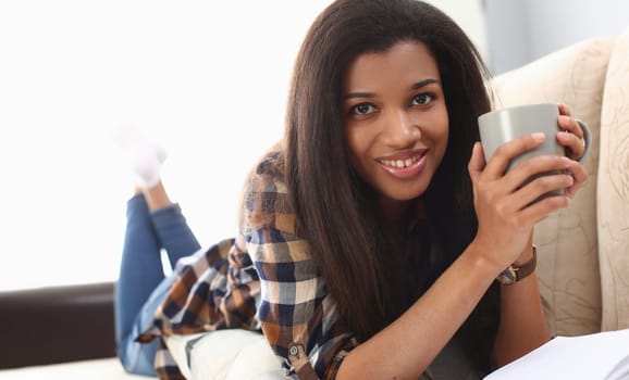Black woman enjoys relaxing with coffee lying on sofa. Home leisure and rest
