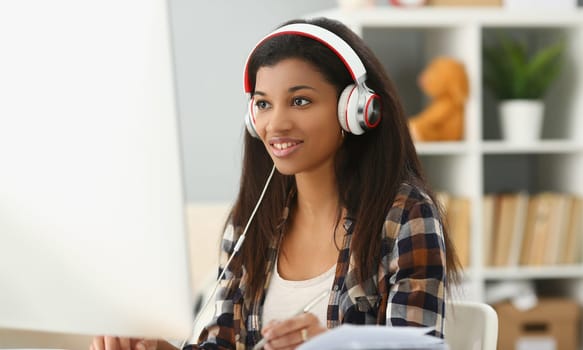 Smiling young black woman in headphones sitting at desk. Woman freelancer working on computer and writing notes in notebook and watching webinar