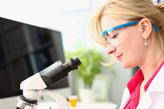 Woman scientist in glasses conducts research through microscope in chemical laboratory. Study of new bacterial viruses and toxic substances