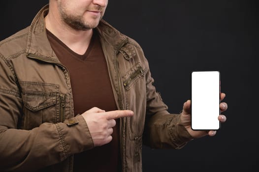 Smiling man with big blank cell phone in hand showing white screen at camera pointing at you, recommending new app or mobile website, banner collage mockup, pointing finger at device.