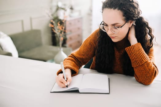 Young woman multitasking: writing in journal and studying for success. Close-up view of woman writing in copybook for studying or making checklist. Focused young woman taking notes in paper notebook.