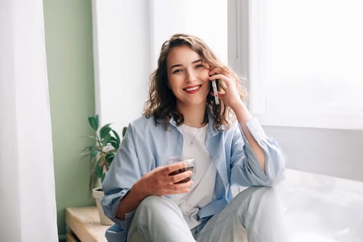 Happy woman chatting on phone with sister or friend over tea on kitchen windowsill. Pleasant conversation with loved ones. Joyful young woman having fun talking on phone while drinking tea at home.